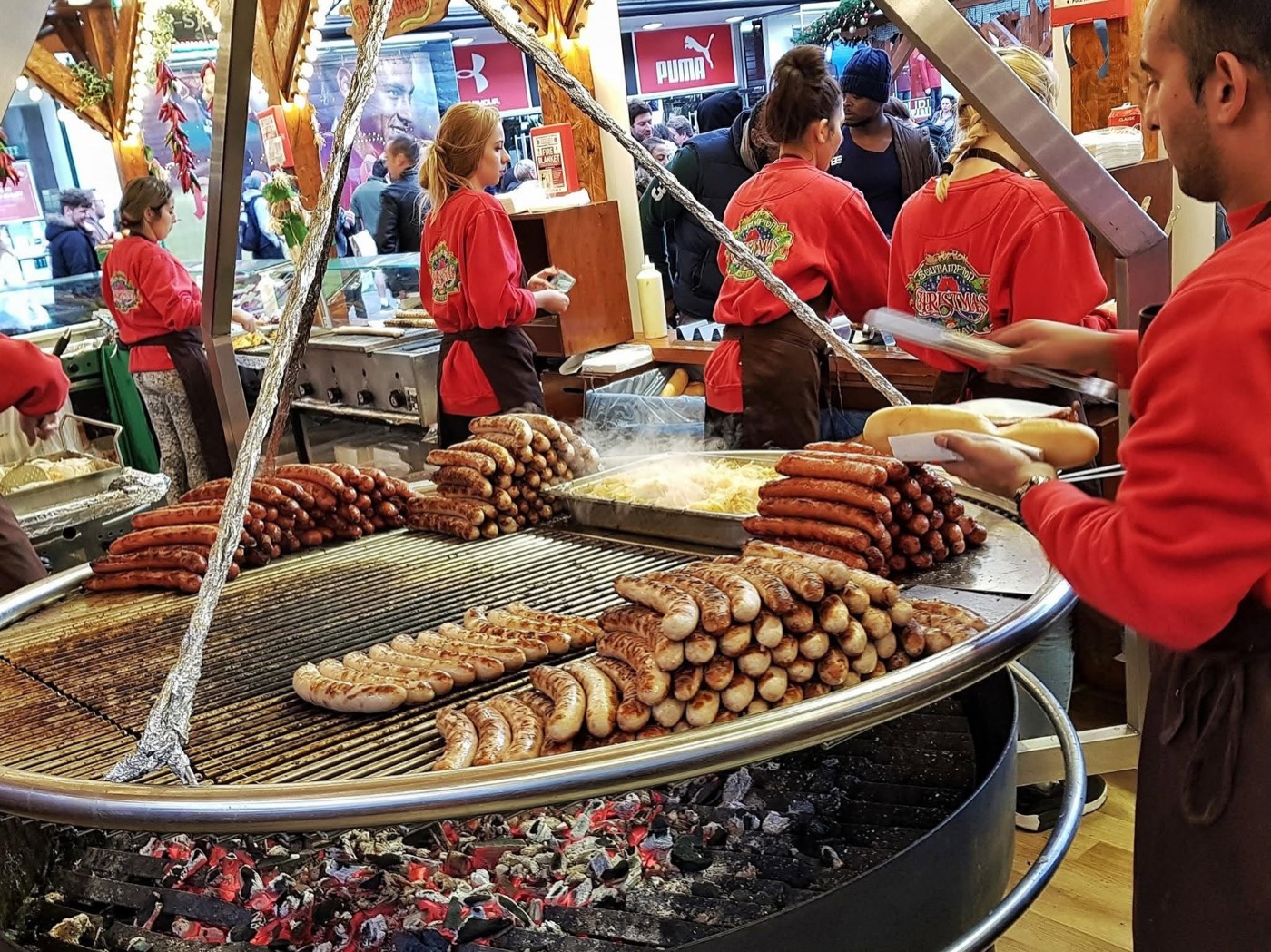 A grill piled high with German bratwurst, staff serving in red aprons