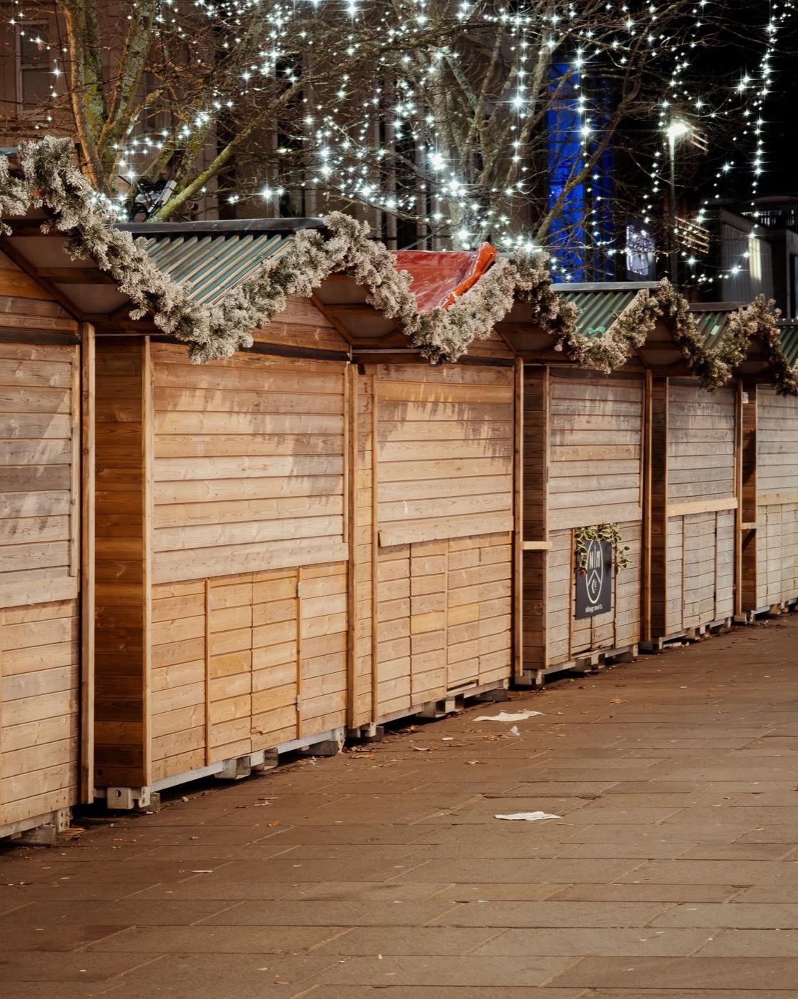 Rows of shuttered wooden market stalls lit by fairy lights at night